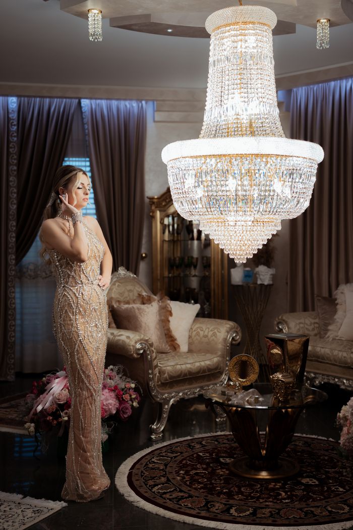 A sophisticated woman in glamorous attire stands beside a grand chandelier in an opulent room.